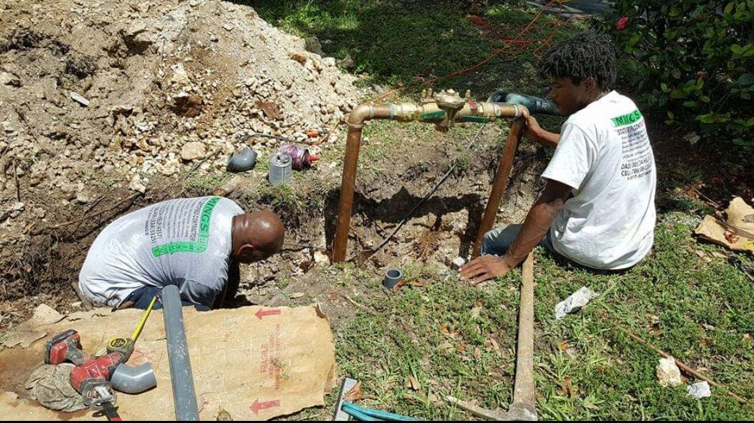 Two people working on plumbing in a dirt excavation on grass. One is in the hole, the other is holding a pipe.