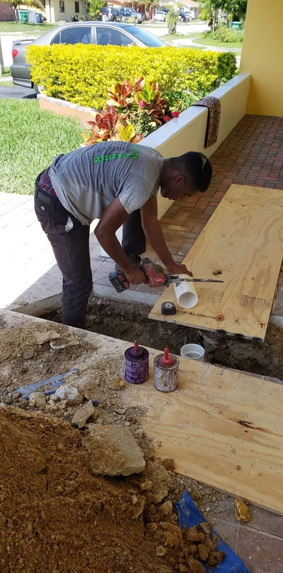 A person is working on plumbing in a trench. They are using a drill and other tools on a piece of wood.