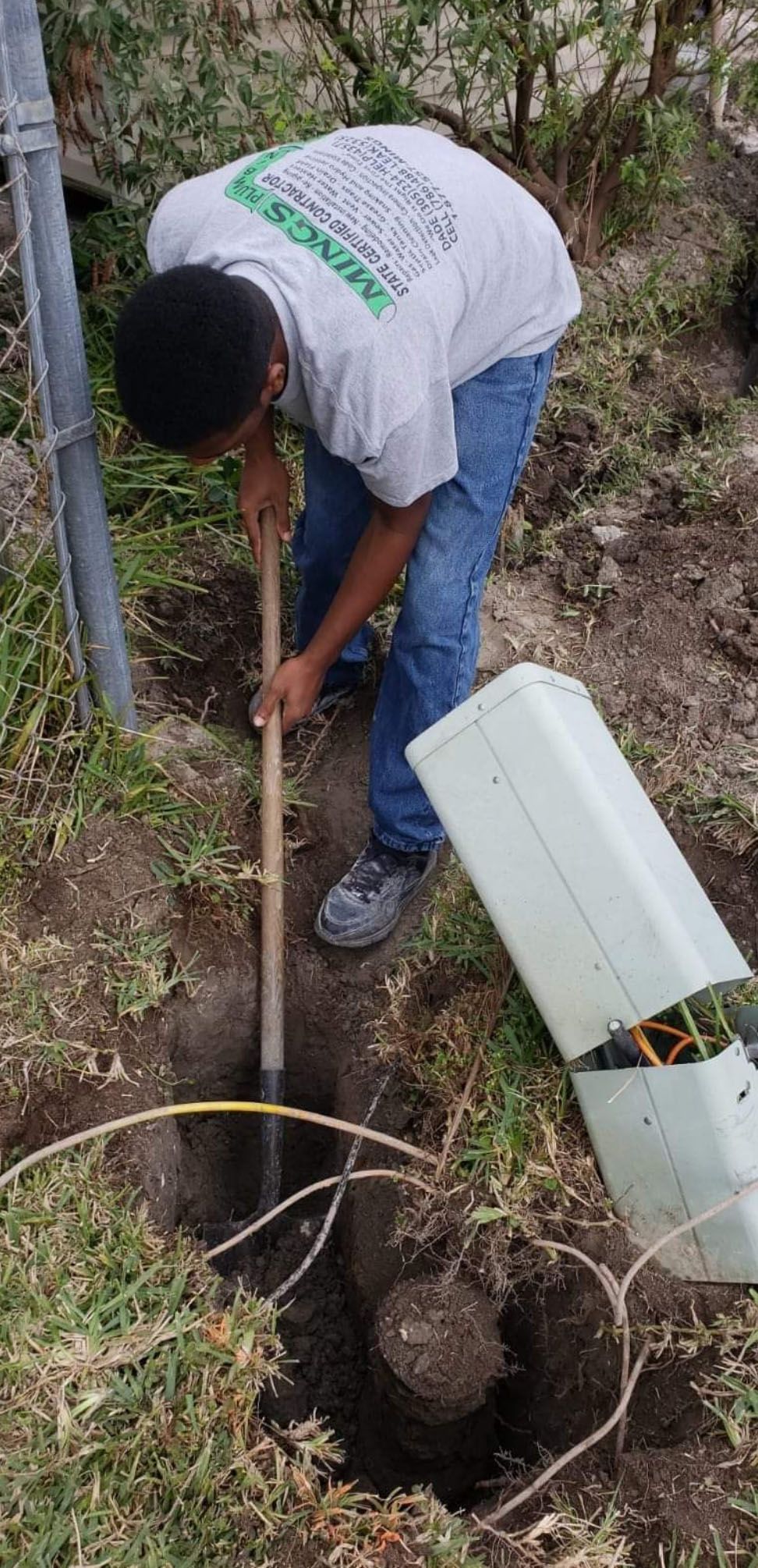 Person digging in a hole with a shovel near a white electrical box in a grassy area.