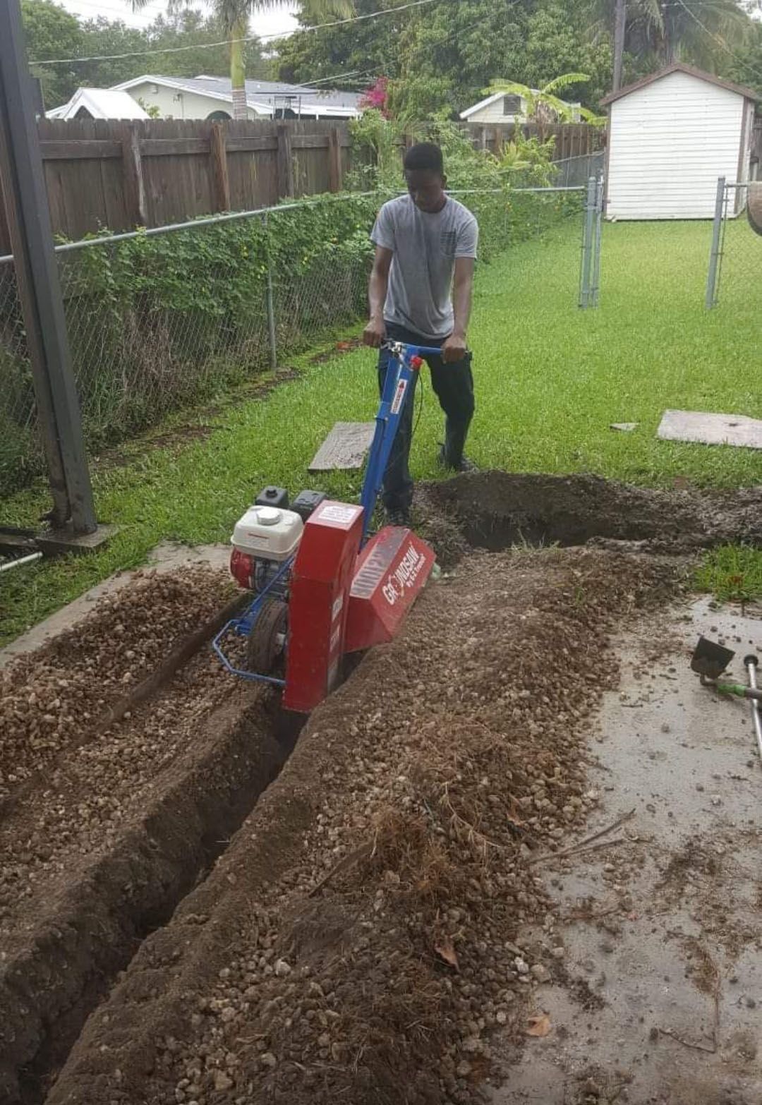Person operating a trenching machine in a backyard, digging a ditch.