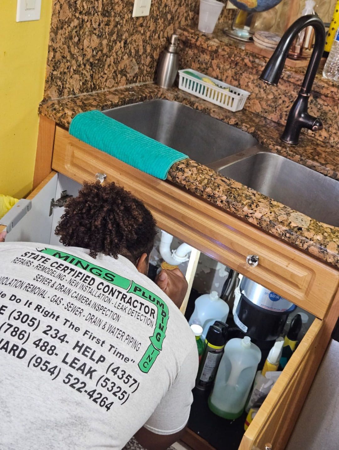 Plumber working under a kitchen sink. Beige cabinets, granite countertop, various cleaning supplies visible.
