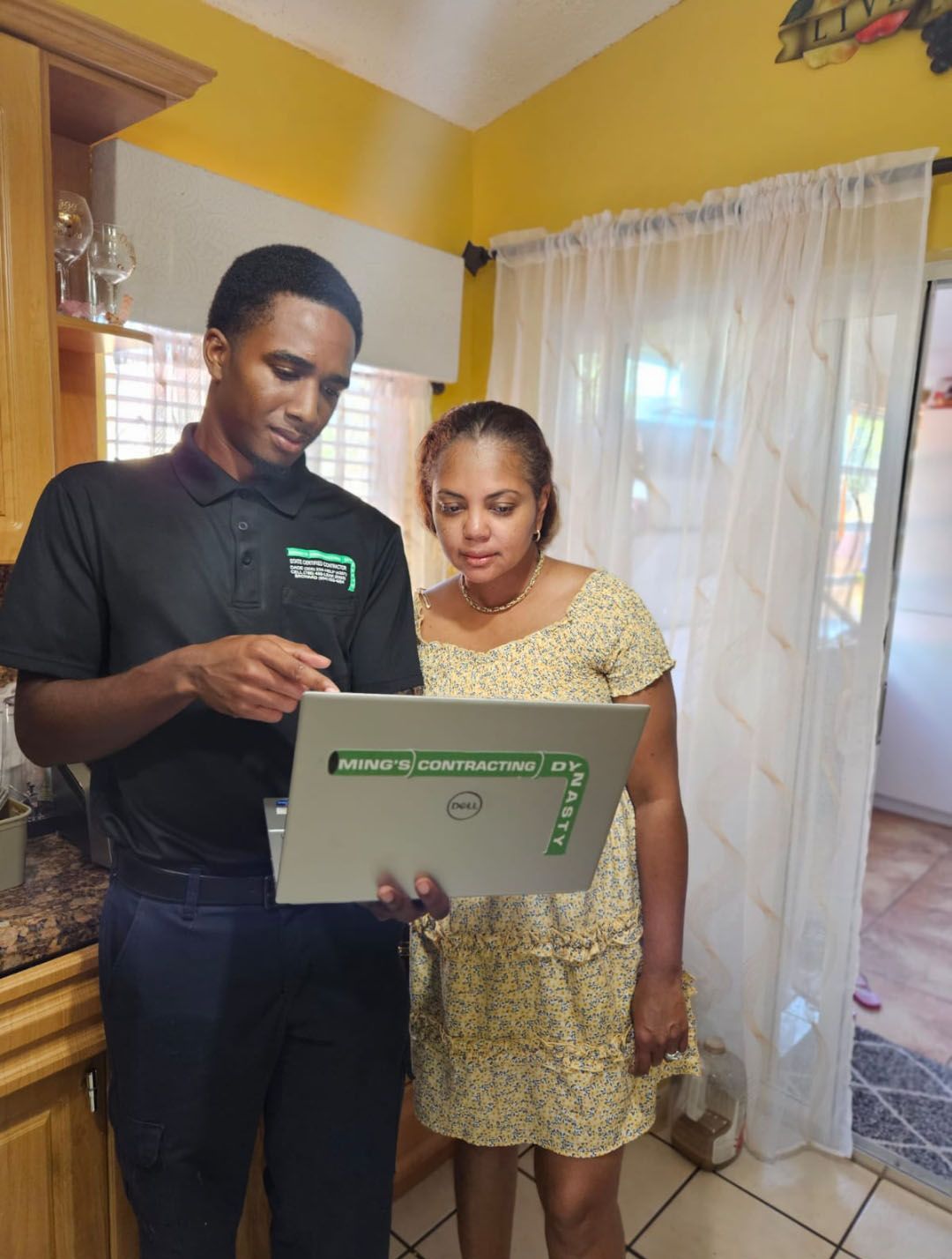 A person in a black polo shirt shows a laptop to another person in a floral dress in a kitchen.