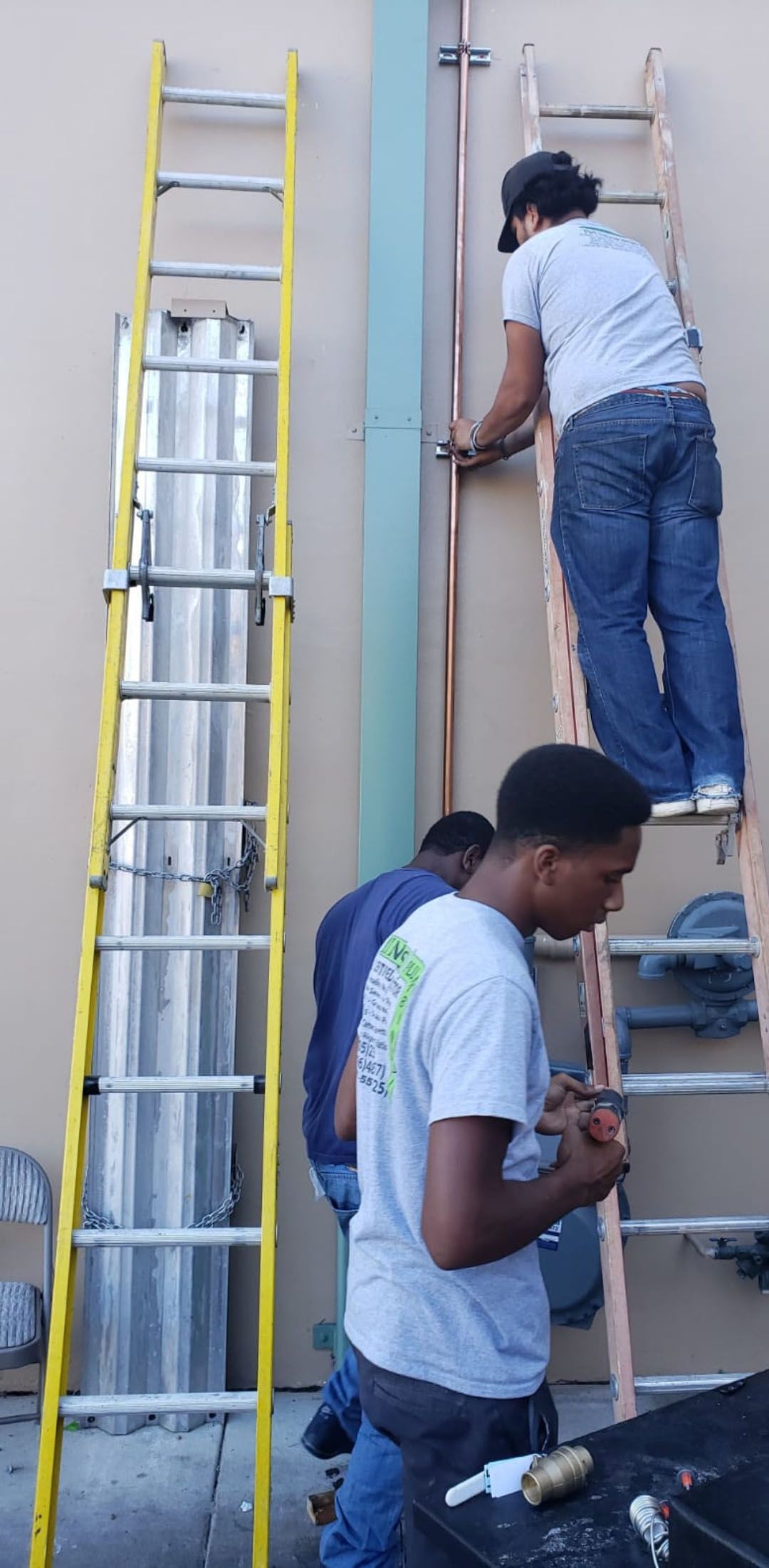 Three workers installing copper piping on a building exterior. Ladders are used.