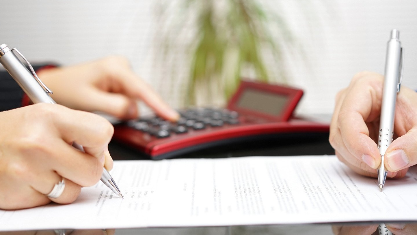 Two people are writing on a piece of paper with a calculator in the background.