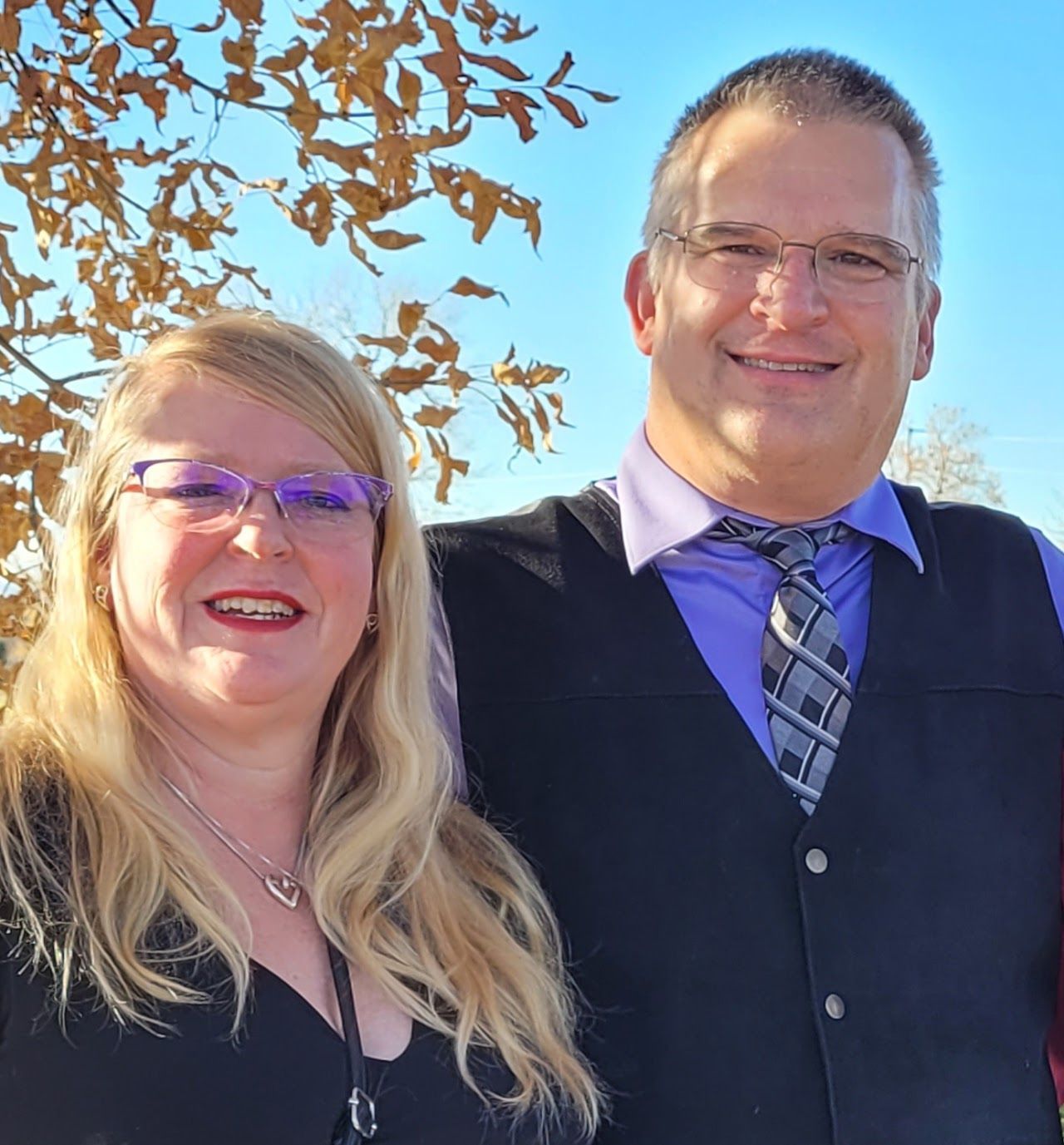 Woman and man smiling, standing close. She has blonde hair and glasses. He wears a vest and tie. Outdoors, blue sky.