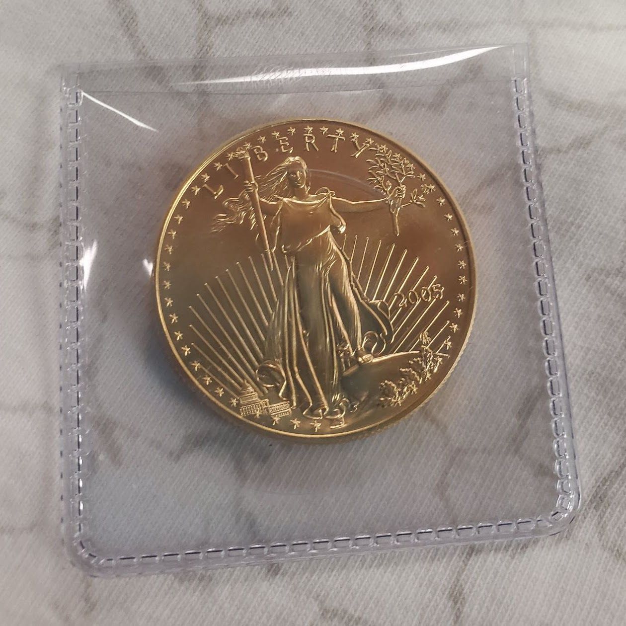 Gold coin in a clear plastic sleeve. The coin features a standing figure, and is golden.