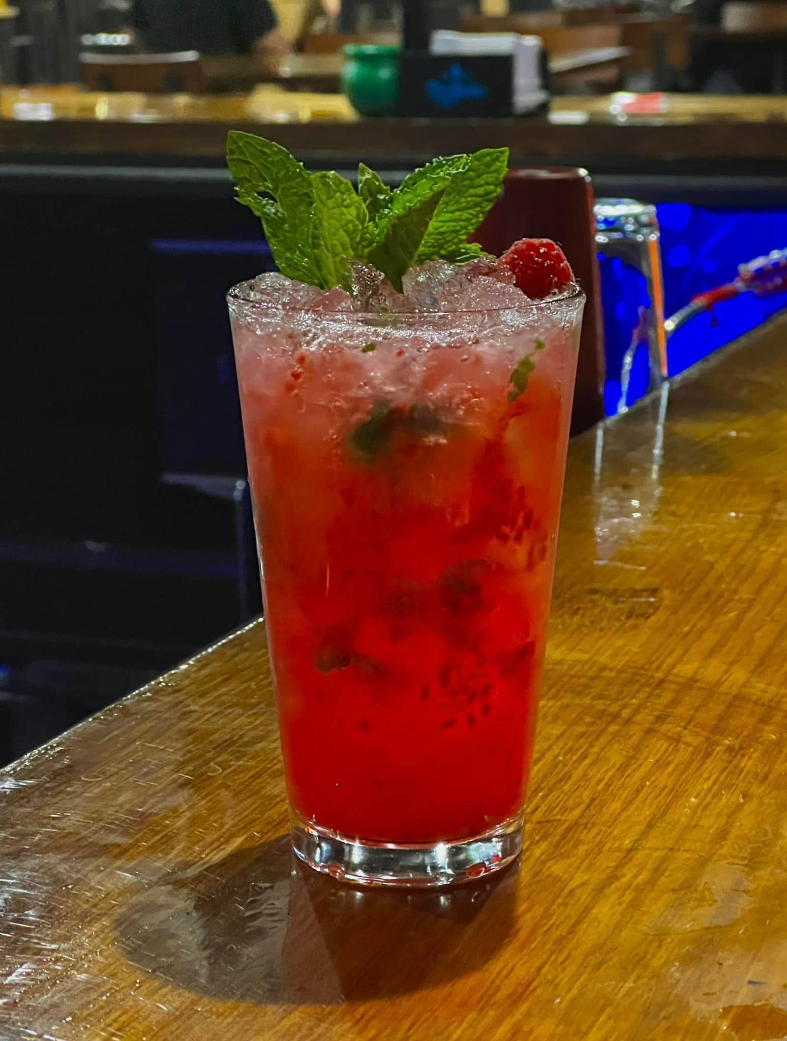 A red drink with mint leaves on top is on a bar.