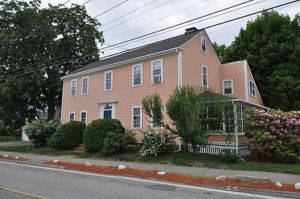 Two-story peach-colored house with multiple windows and a porch. Green shrubs and trees surround the building.