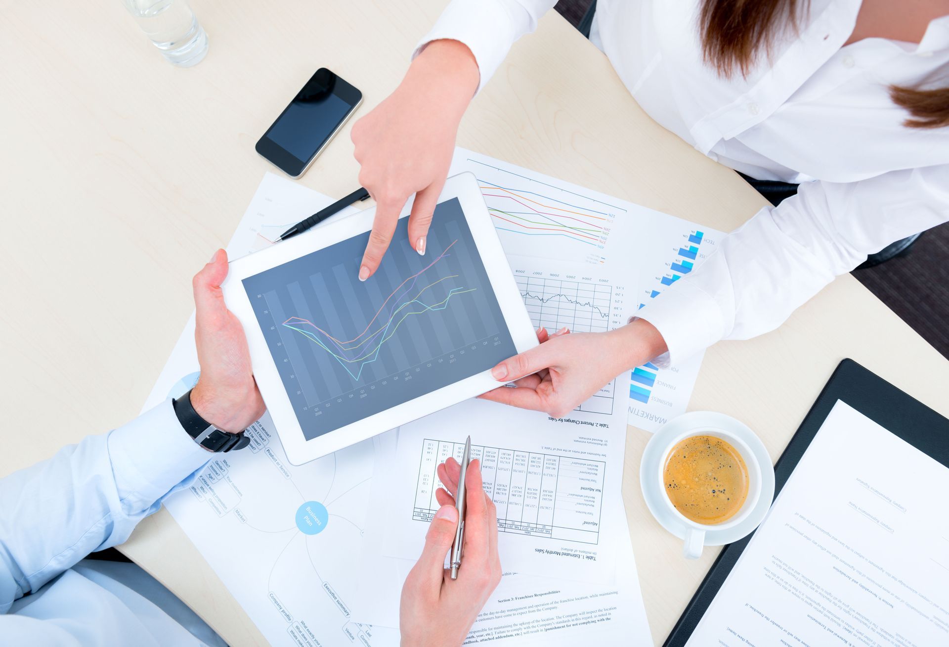 Hands reviewing charts and a tablet at a bright office desk with coffee and phone.
