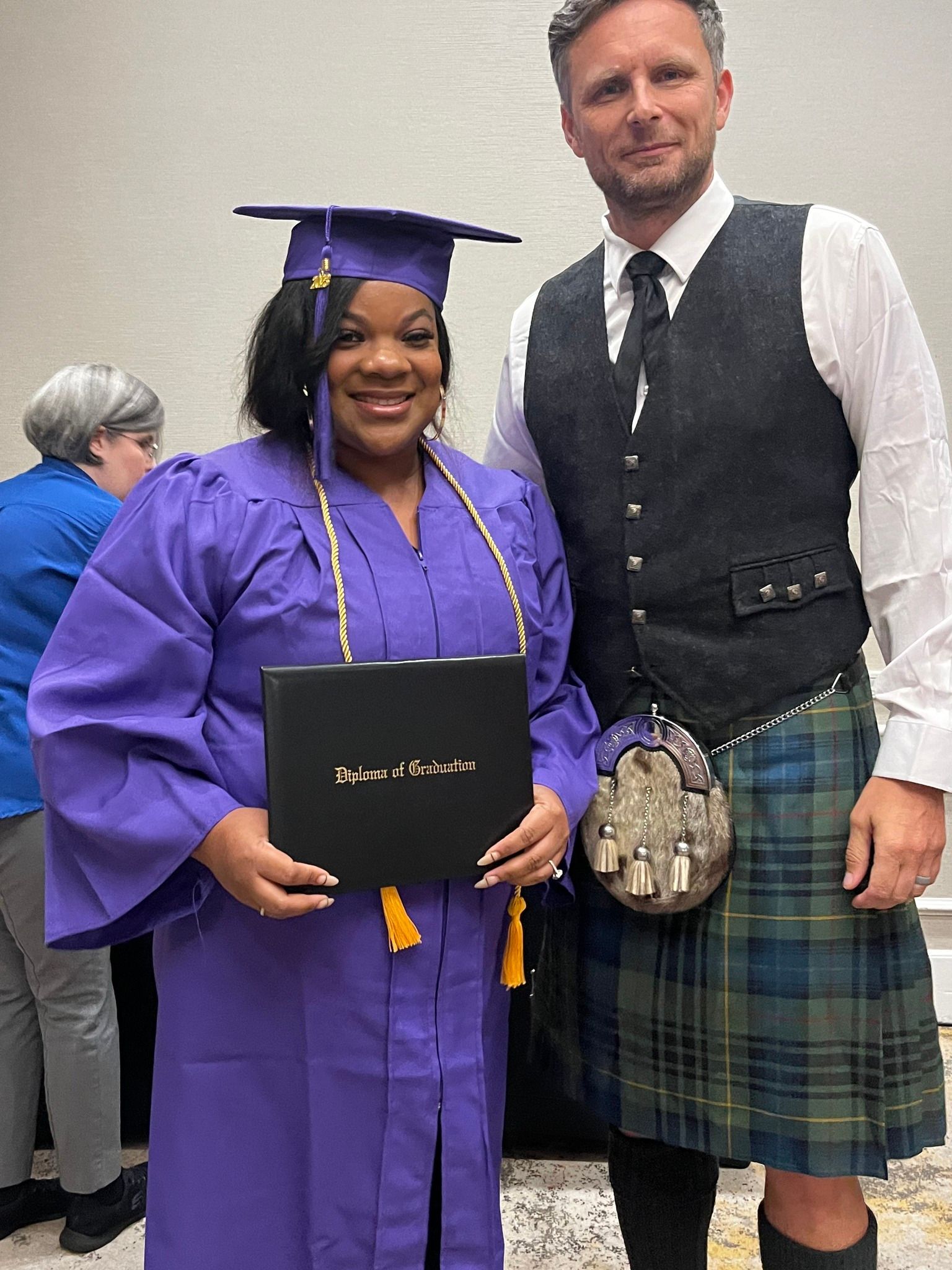 A man in a kilt is standing next to a woman in a purple cap and gown holding a diploma.