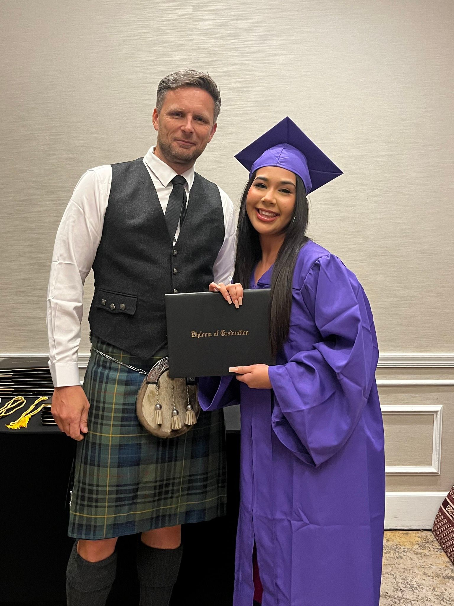 A man in a kilt is standing next to a woman in a graduation cap and gown holding a diploma.