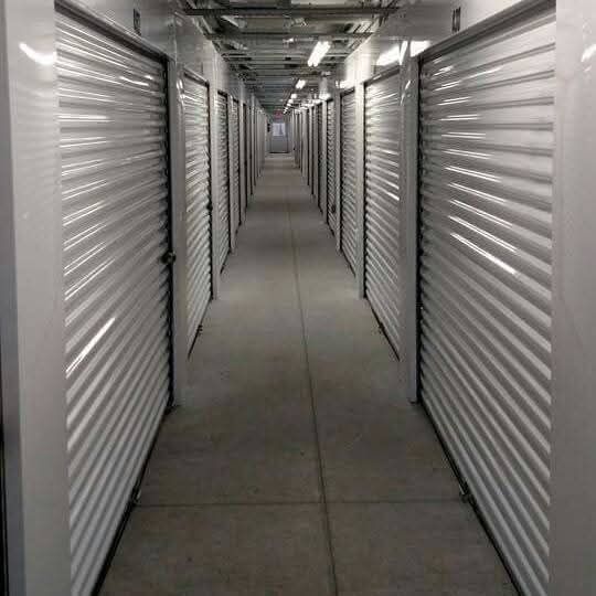 Interior of a storage facility hallway. White metal storage unit doors line both sides, leading toward a distant vanishing point.