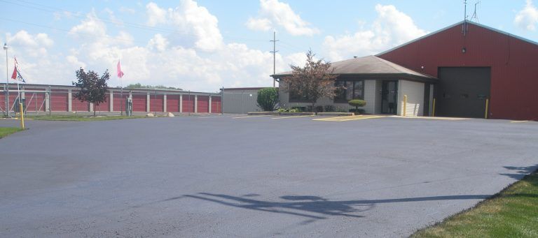 Paved parking area in front of a storage facility with red and beige buildings under a cloudy sky.
