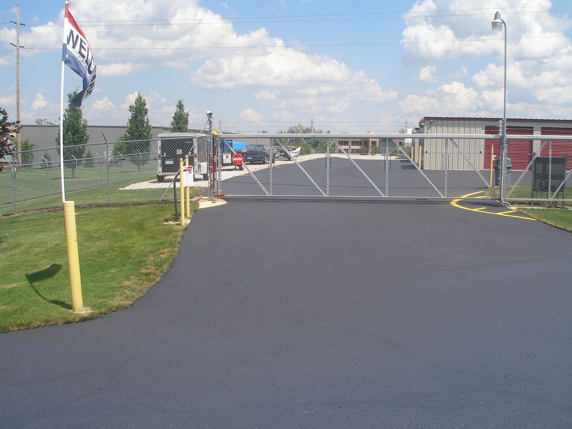 Asphalt driveway leading to a gated storage facility entrance with flag, blue sky.