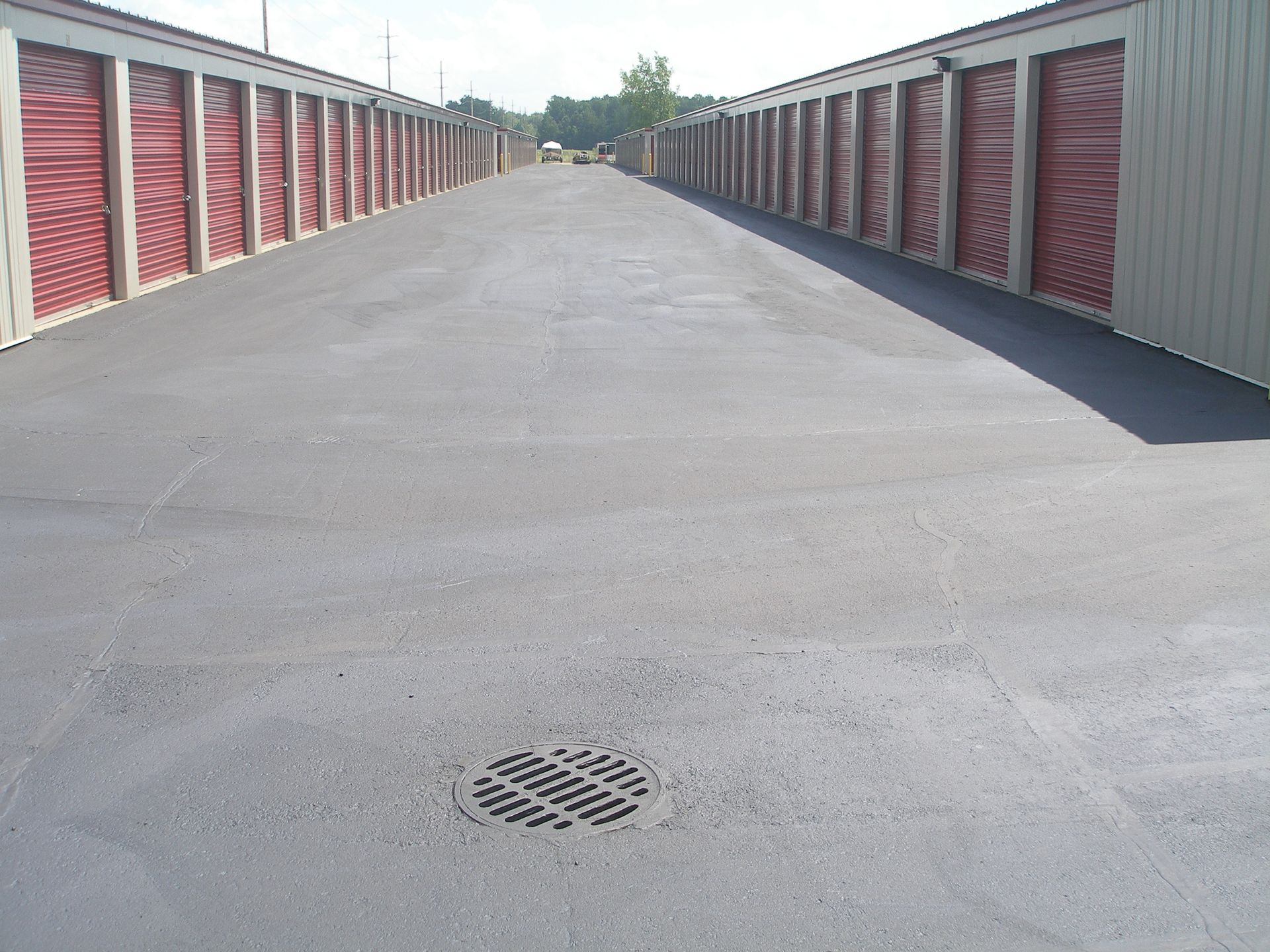 Rows of storage units with red doors flank a gray asphalt drive. A drain is in the foreground.