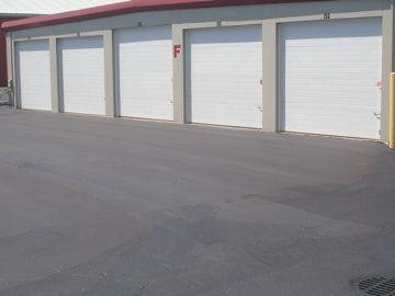 Storage units with white doors, in a row, with a black asphalt parking area in front of them.