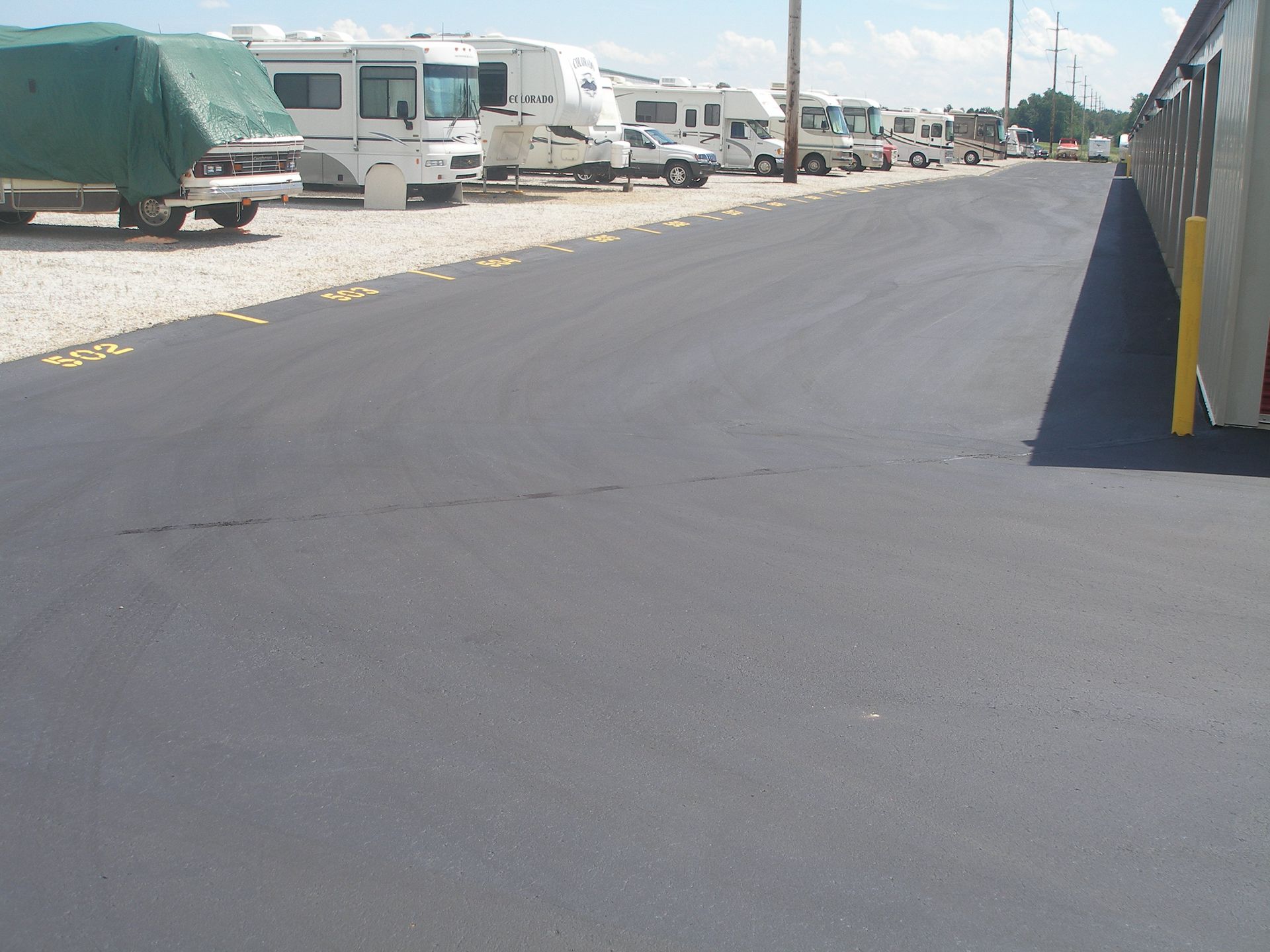 Asphalt road in RV park, RVs parked on gravel, sunny day.