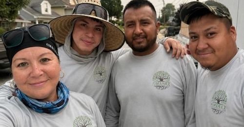 Four people in gray shirts and hats pose outdoors, smiling at the camera.