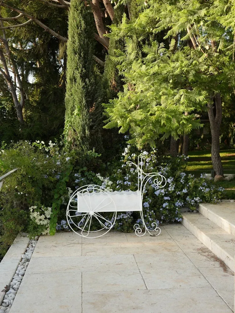 White ornate bench in a garden with trees and greenery beside a stone path