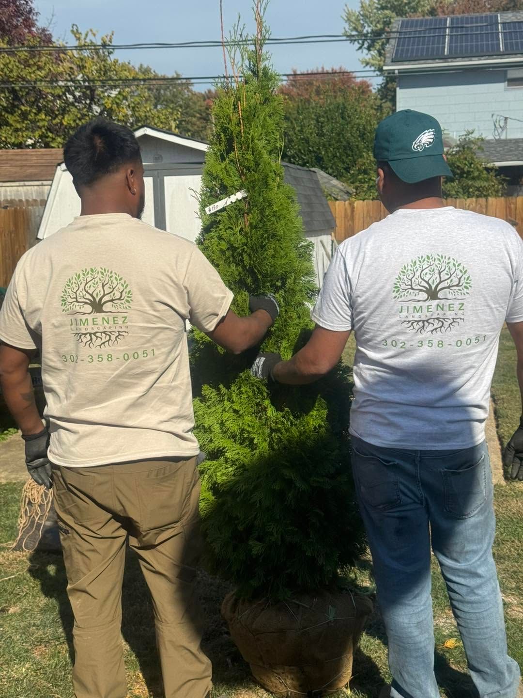 Two people in tan and white shirts stand outdoors beside a trimmed evergreen shrub.