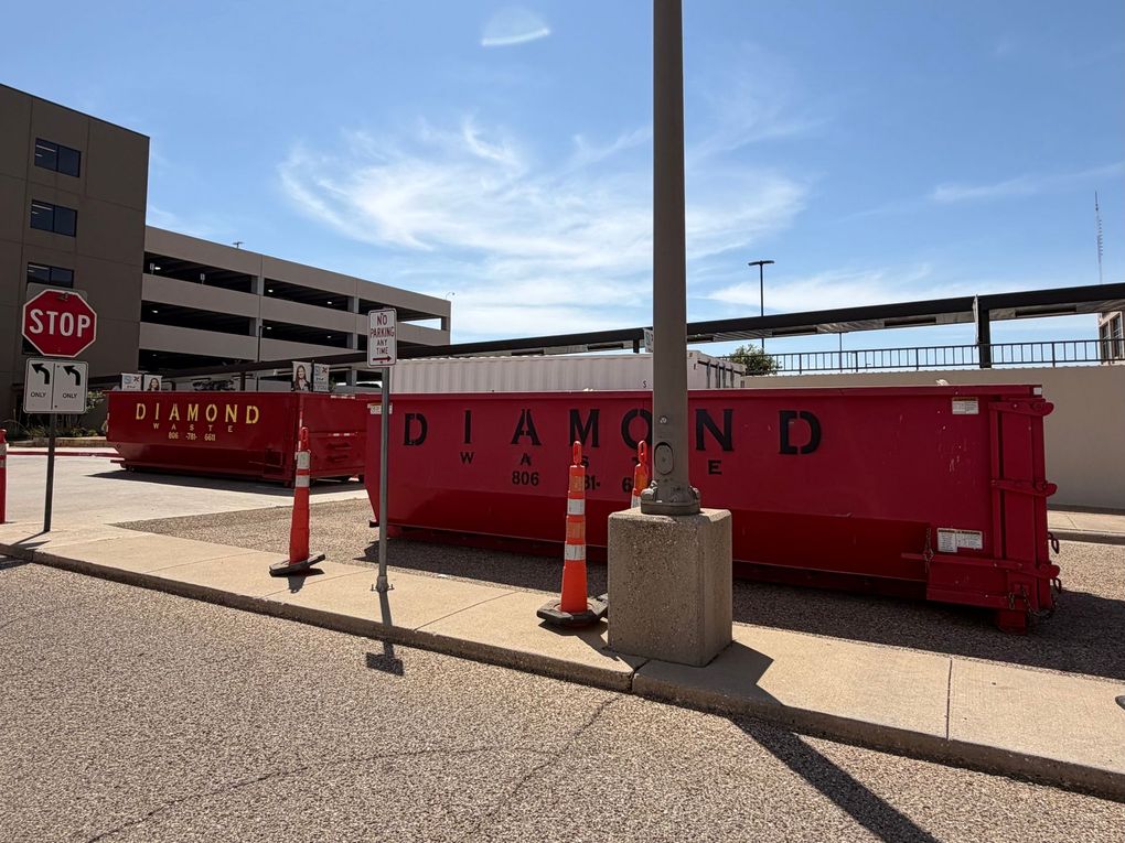 Two red Diamond Waste dumpsters at a commercial property, surrounded by traffic cones and parking structures.