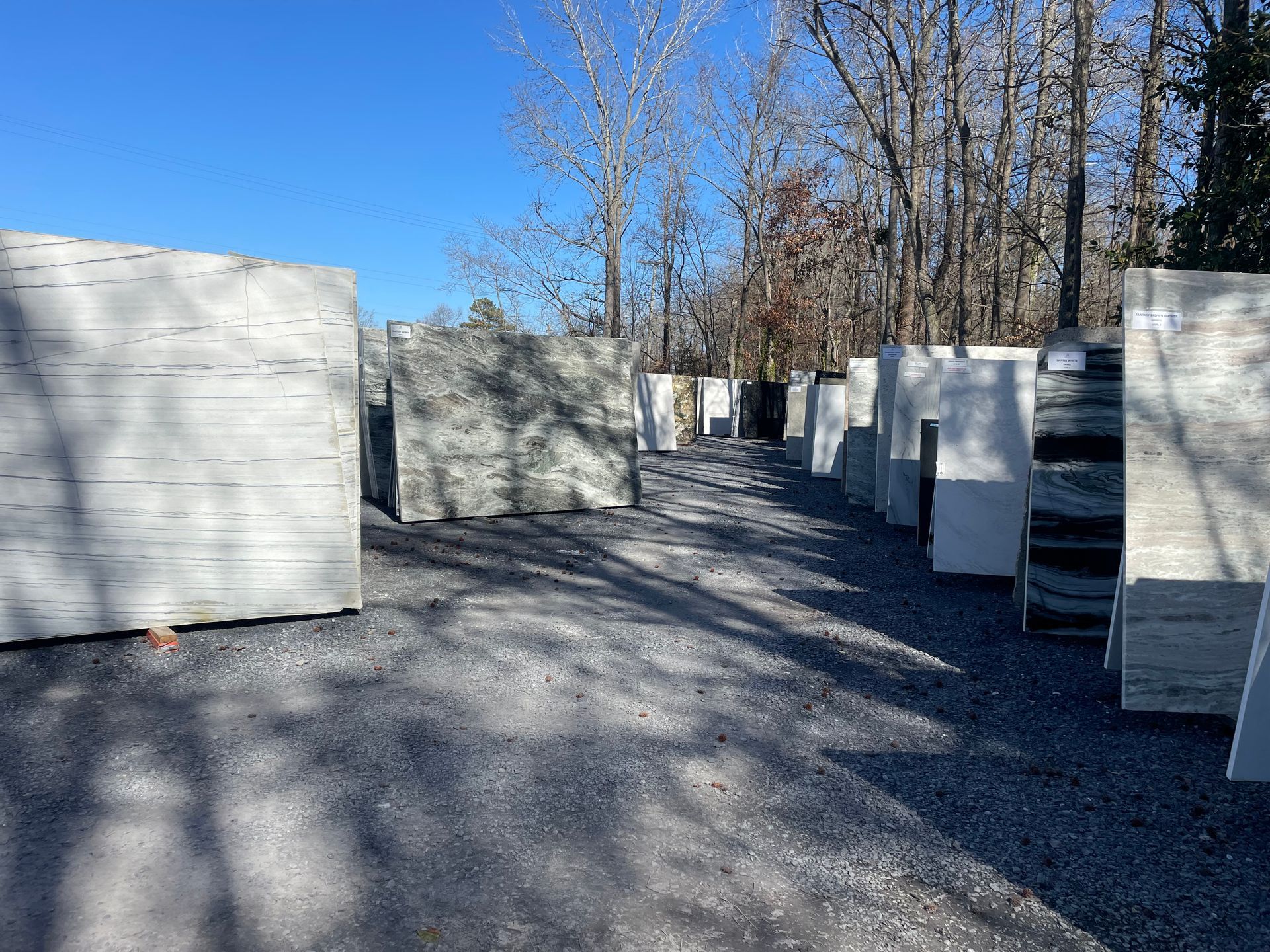 Rows of large gray stone slabs outdoors on gravel, under a blue sky, with trees in the background.