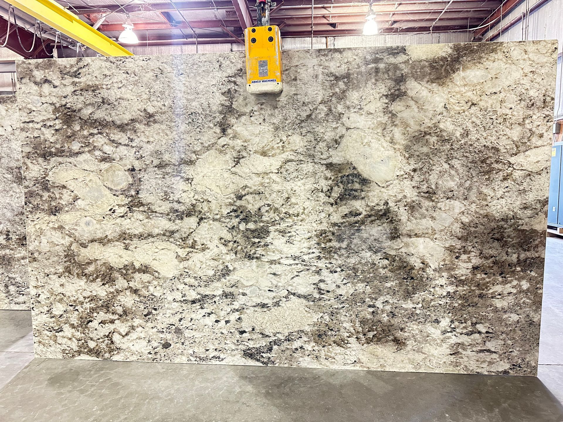 Large granite slab with cream, brown, and gray patterns, held by a yellow lifting device in a warehouse.