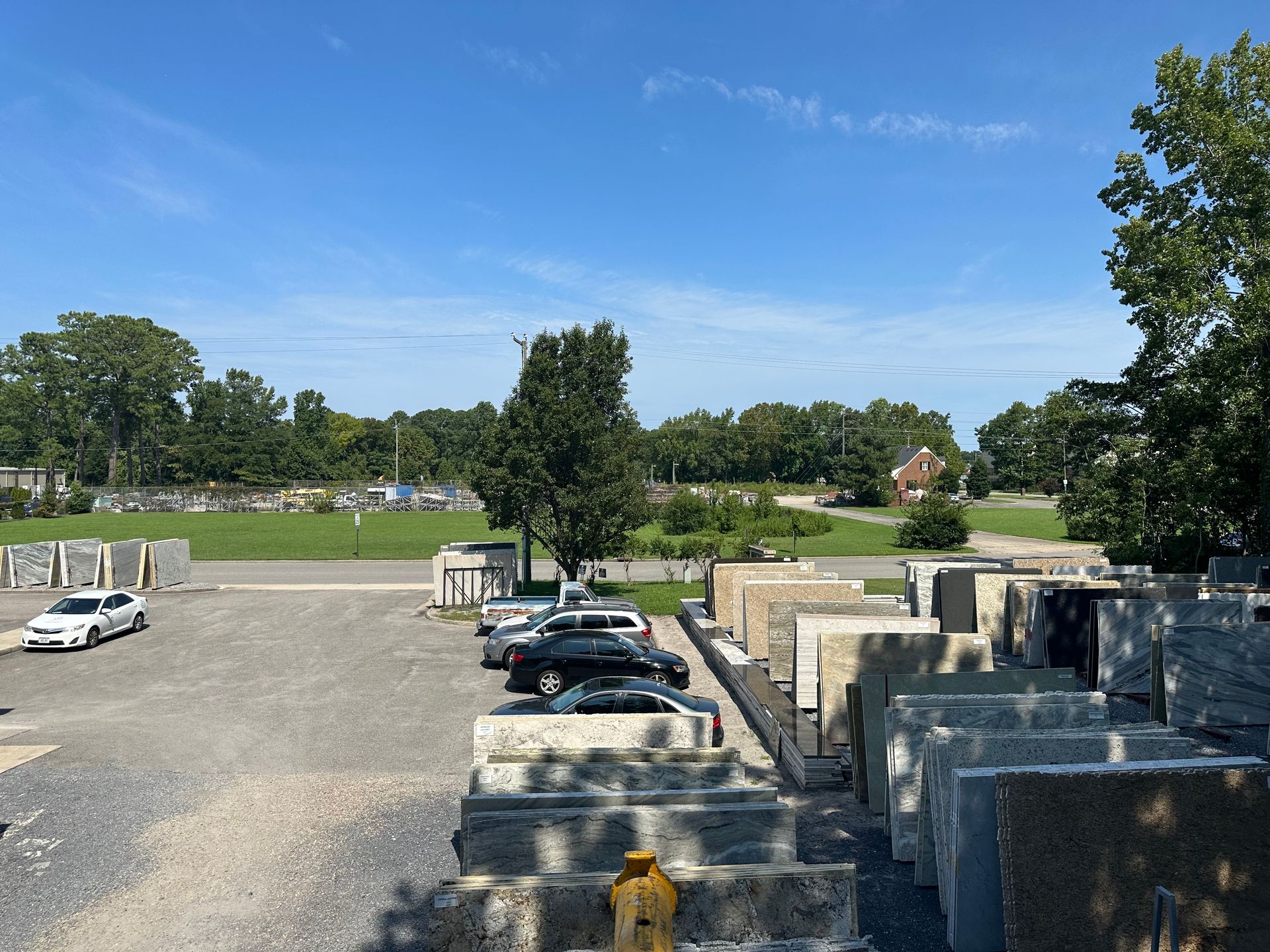 Gravel lot with stacked stone slabs, cars, trees, and cemetery under a blue sky.