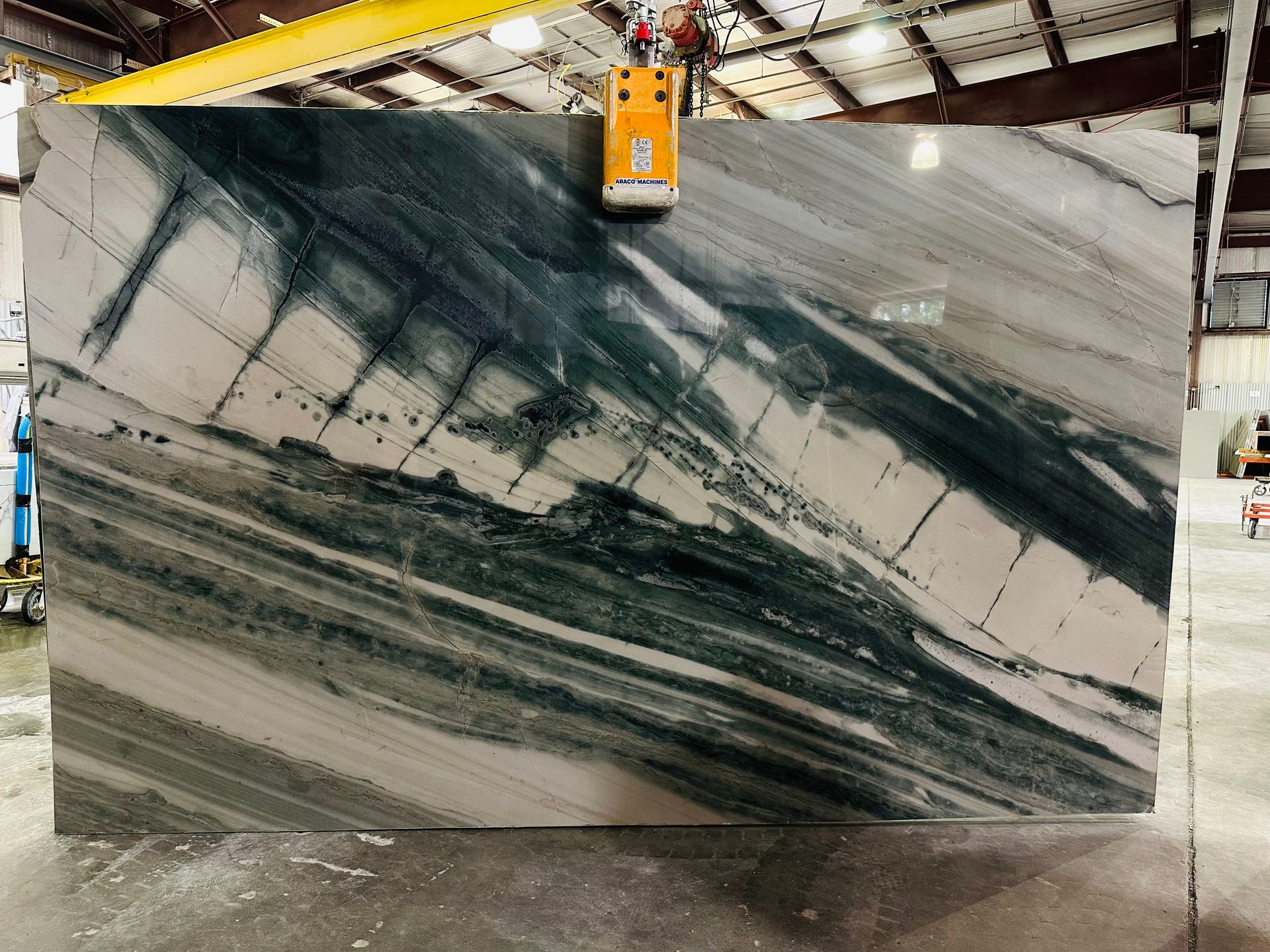 Large marble slab with white and green veining, held by an overhead crane in a warehouse.