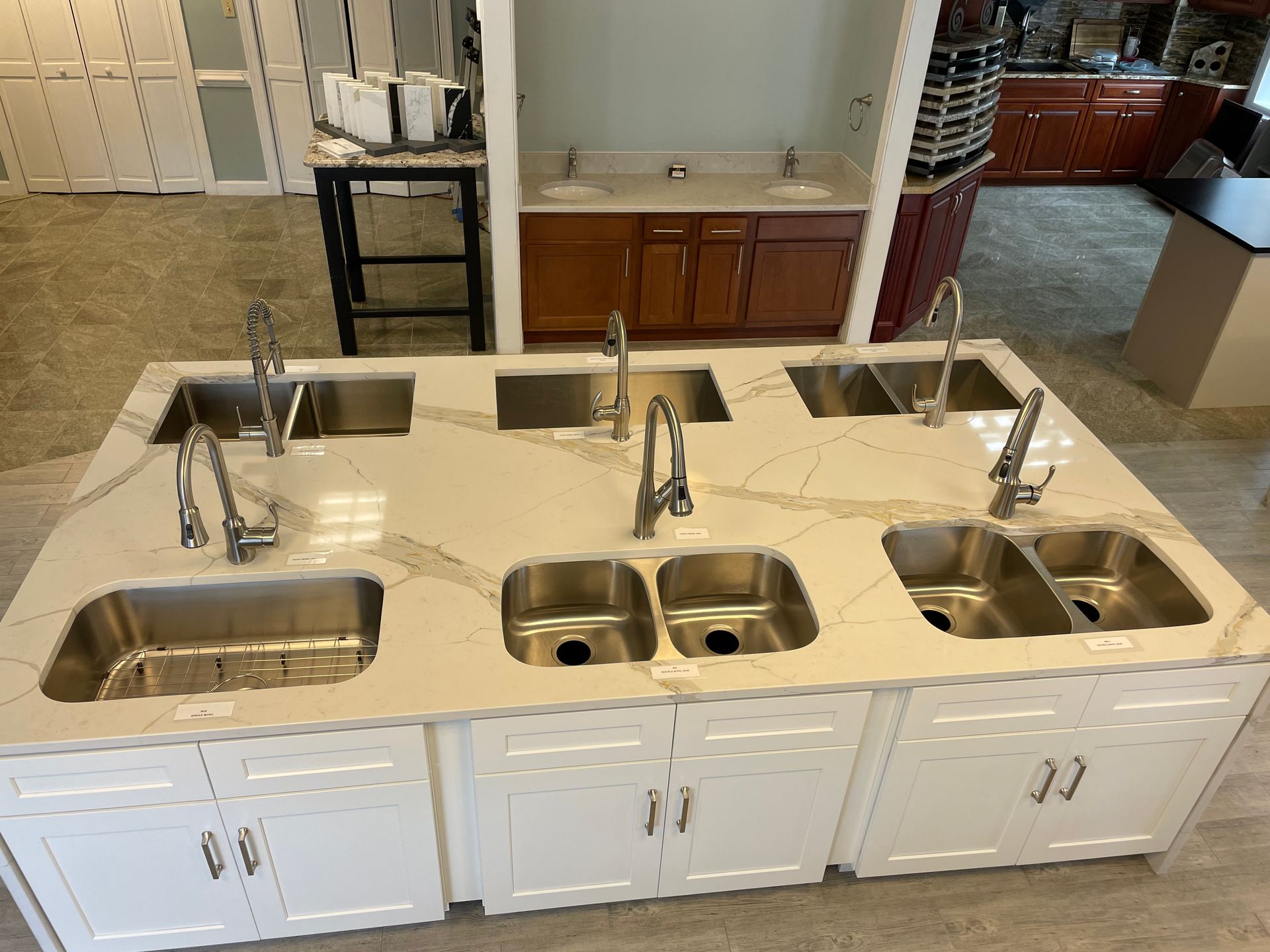 Kitchen island with six sinks and faucets on display in a showroom. Cabinets are white, countertop is light with gray veins.