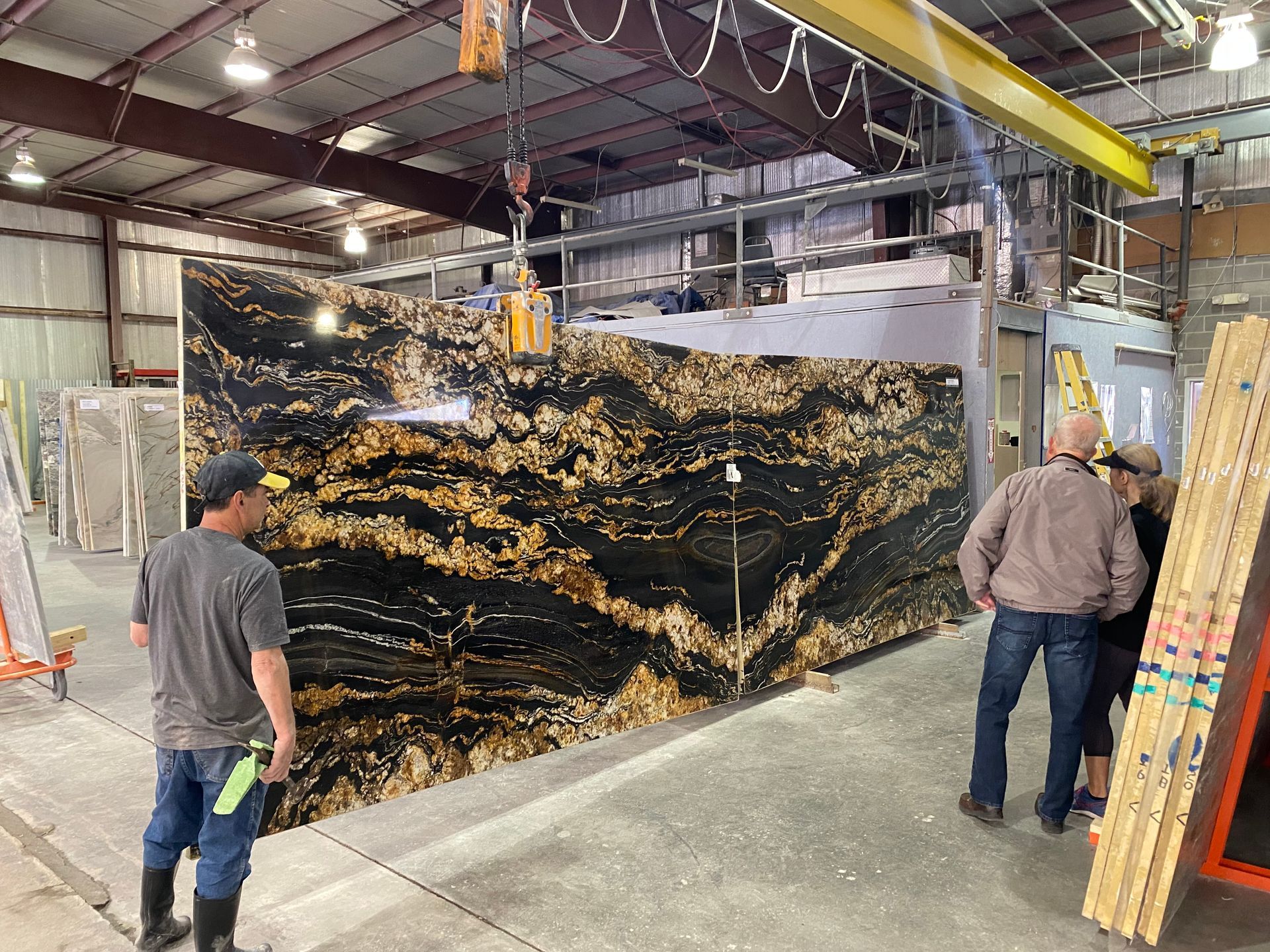 People inspecting large slab of granite in a warehouse. A crane is overhead.