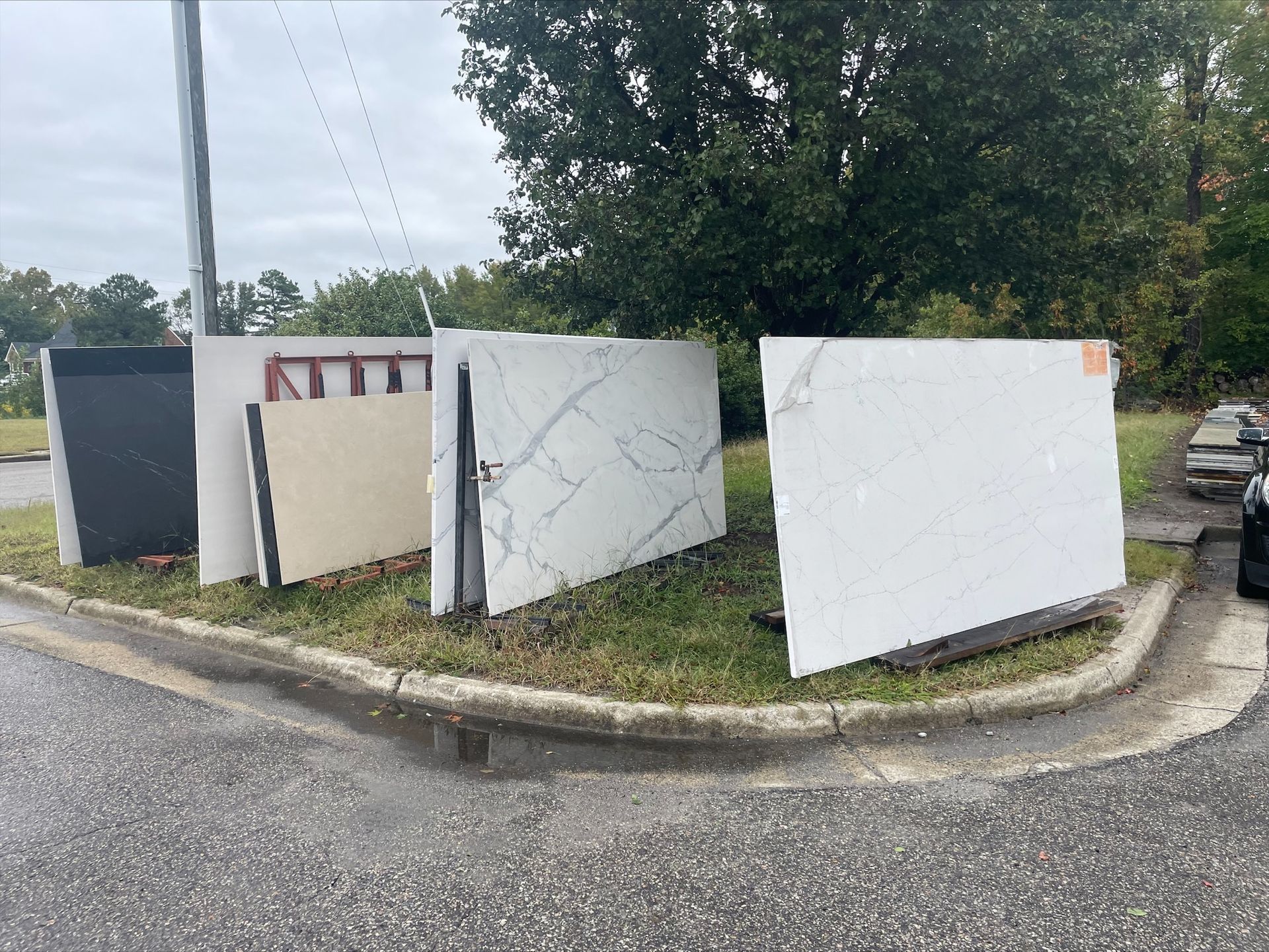 Stone slabs displayed outdoors on a grassy curb in front of trees and a road.