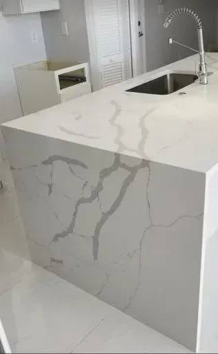 White kitchen island with grey veining, stainless steel sink, faucet, and cabinet visible.