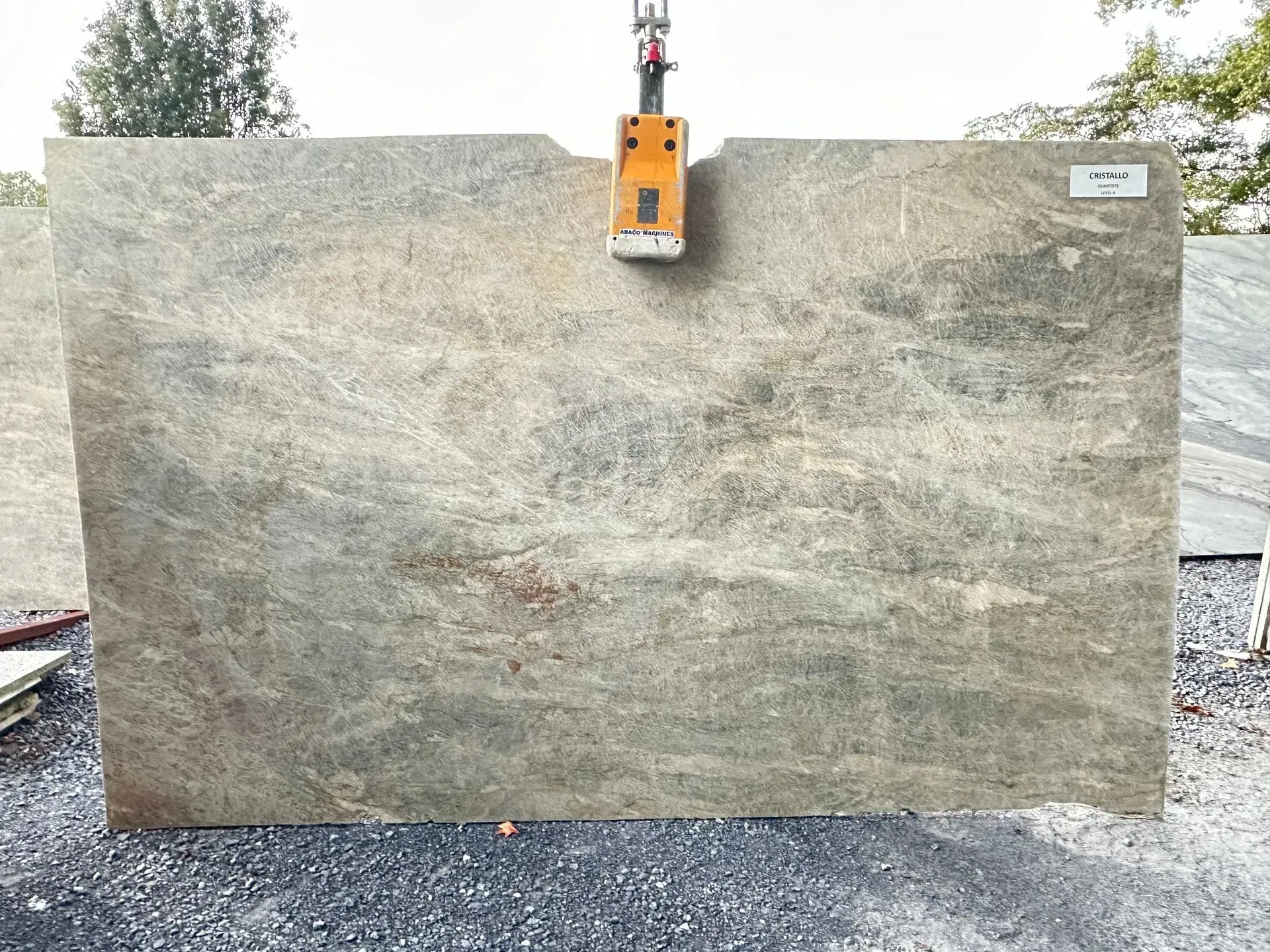 Large, rectangular granite slab, light gray with brown veining, suspended by an orange clamp.