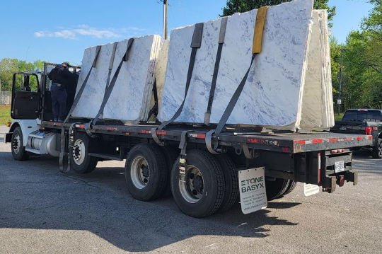 Truck transporting large slabs of light-colored stone, secured with straps. 