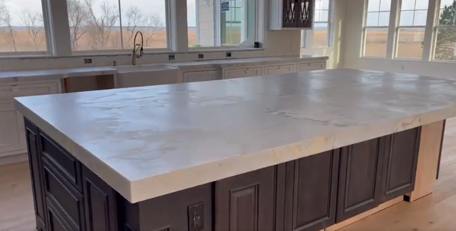 Large kitchen island with light gray countertop, dark cabinets, and surrounding windows.