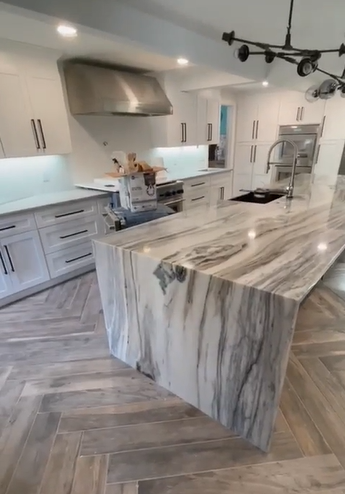 A modern kitchen with a large marble island, white cabinets, stainless steel appliances, and a herringbone floor.