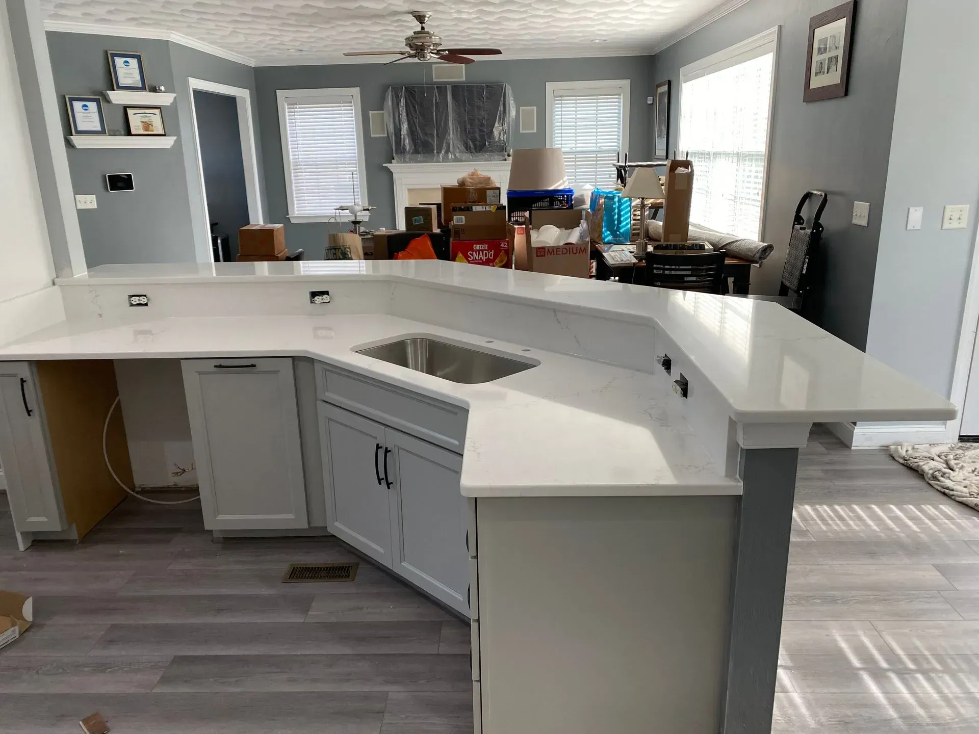 Kitchen island with white countertop, cabinets, and a sink. Gray flooring. Unpacked boxes in background.
