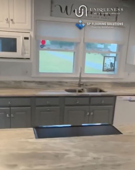 Gray kitchen cabinets with light countertops and a sink in front of a window.