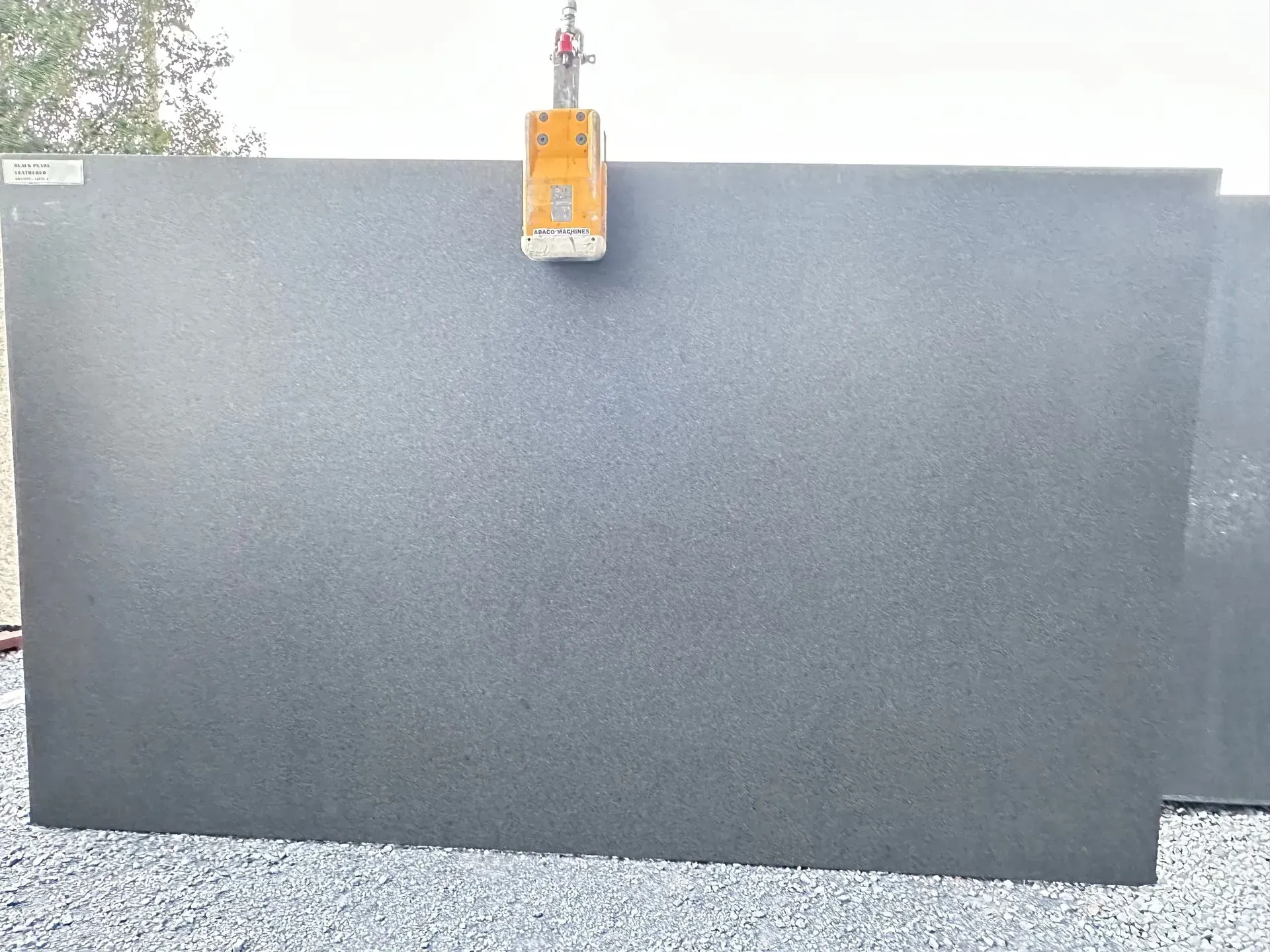 Large gray granite slab being lifted by a yellow crane. Outdoors on gravel.