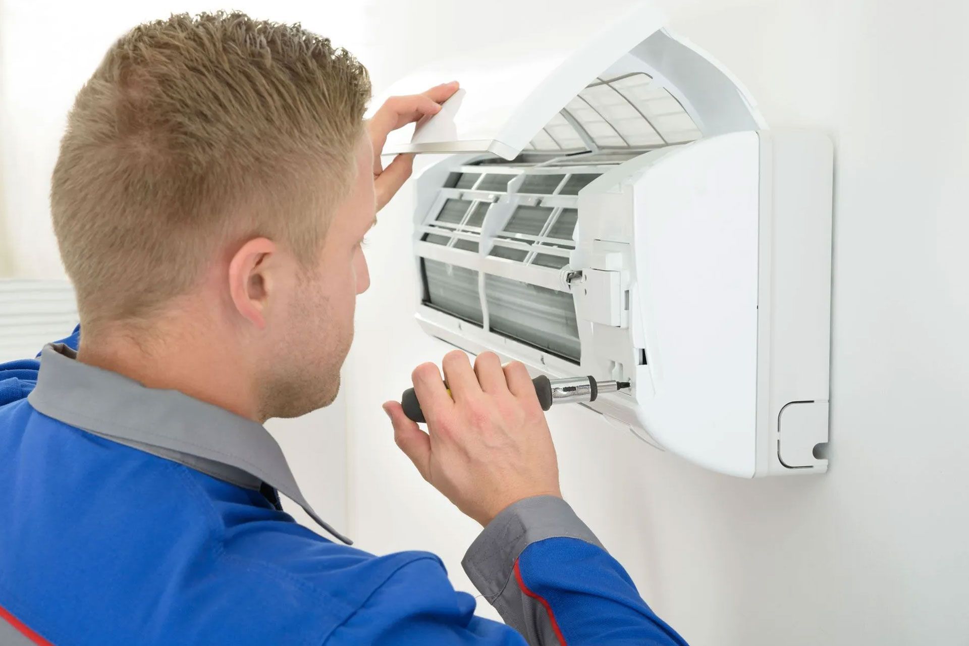 A technician in a blue uniform uses a screwdriver to repair a white wall-mounted air conditioning unit.