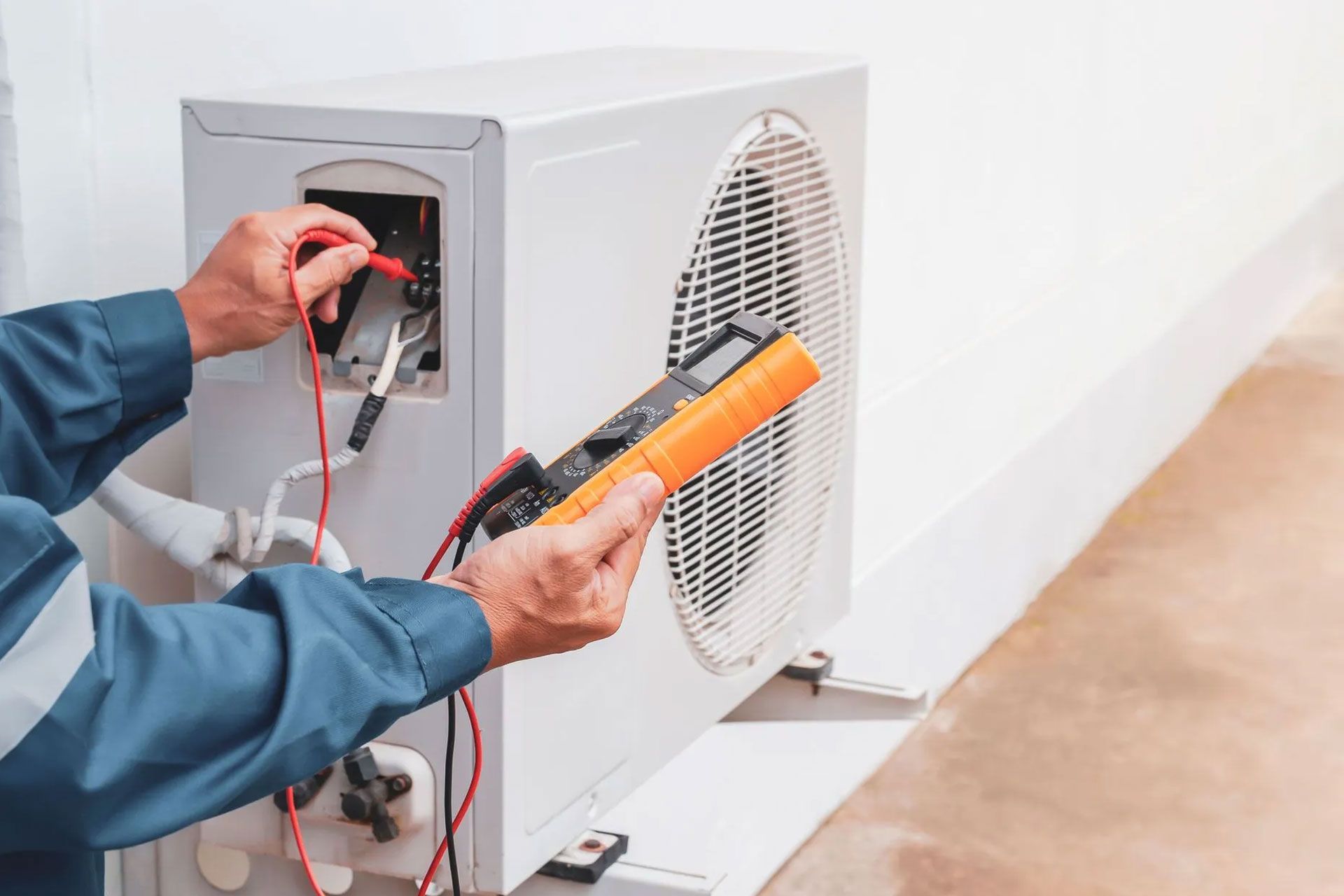 A technician in a blue uniform uses a multimeter to test the electrical connections of an outdoor air conditioning unit.