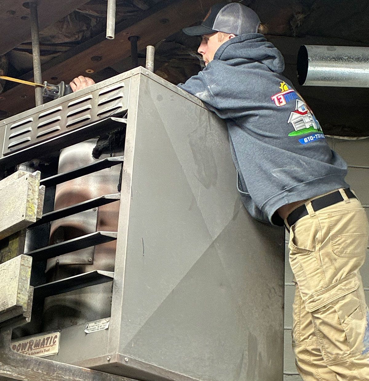 A technician in a gray hoodie and tan pants works on a gray industrial heater mounted in an unfinished interior space.