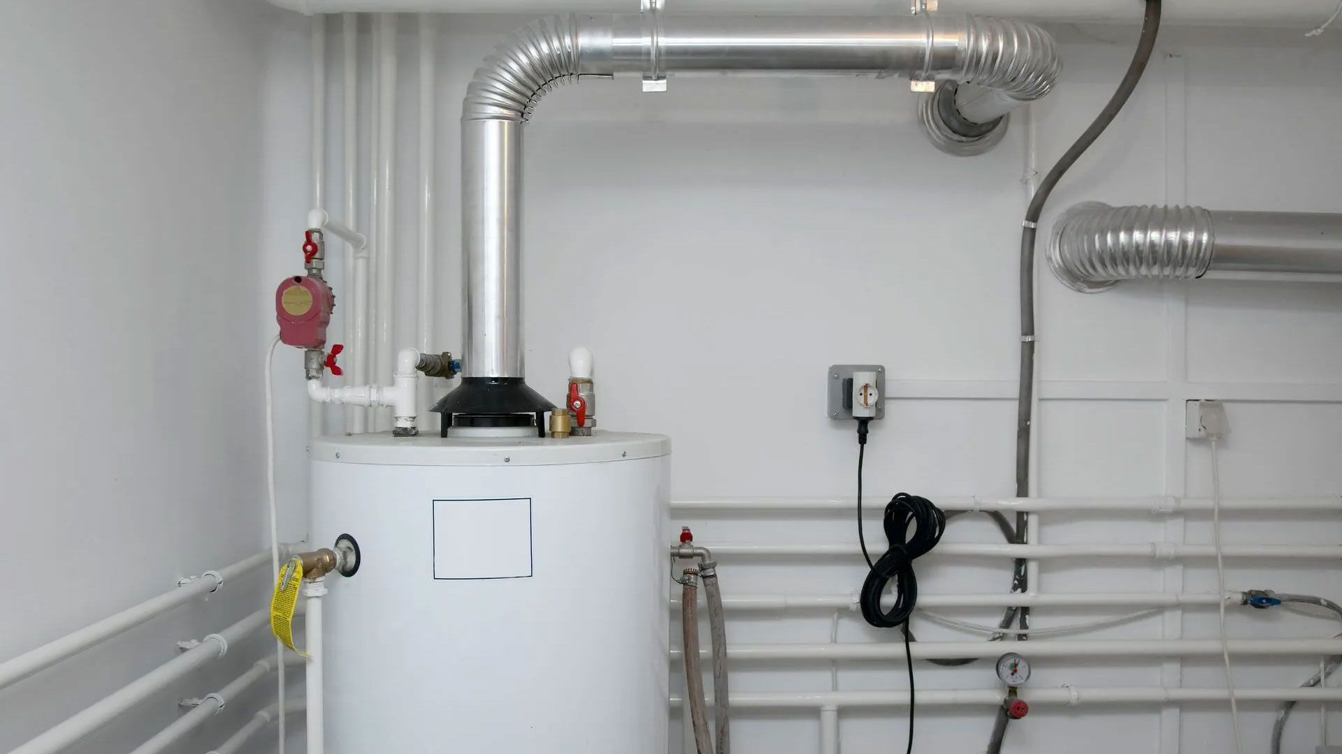 A white water heater stands against a wall with exposed metal venting pipes, electrical wiring, and plumbing.