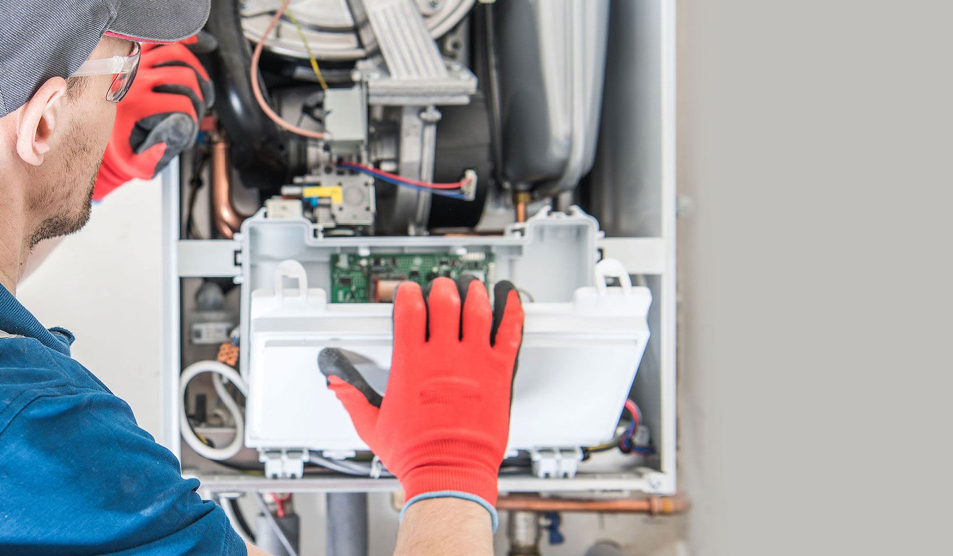 A technician in a blue shirt and red work gloves repairs a wall-mounted gas boiler.