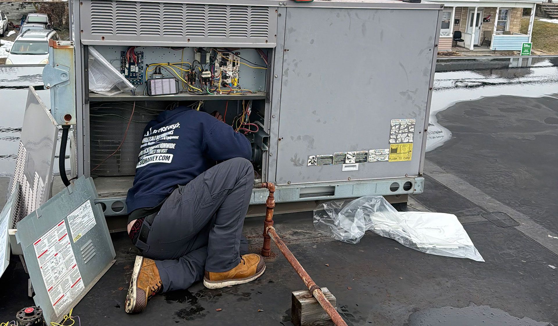A technician in a blue uniform kneels to repair an open rooftop HVAC unit in a parking lot.