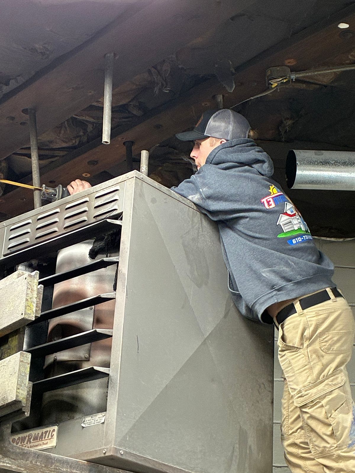 A person wearing a grey hoodie and khaki pants leans against a large, grey industrial heater mounted in a workshop.