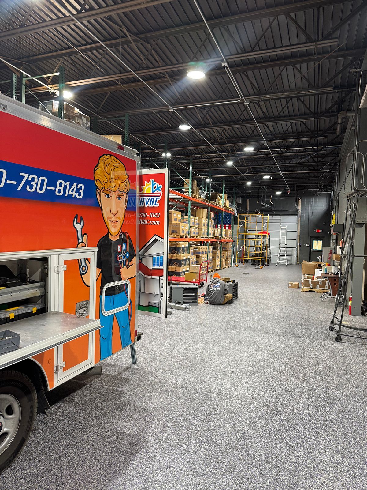 A service van featuring a cartoon technician stands in a warehouse with grey speckled flooring and metal storage racks.