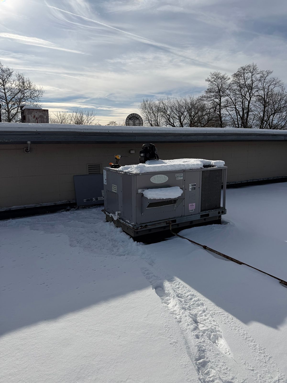 A rooftop HVAC unit sits on a flat roof covered in snow under a bright, partly cloudy sky.