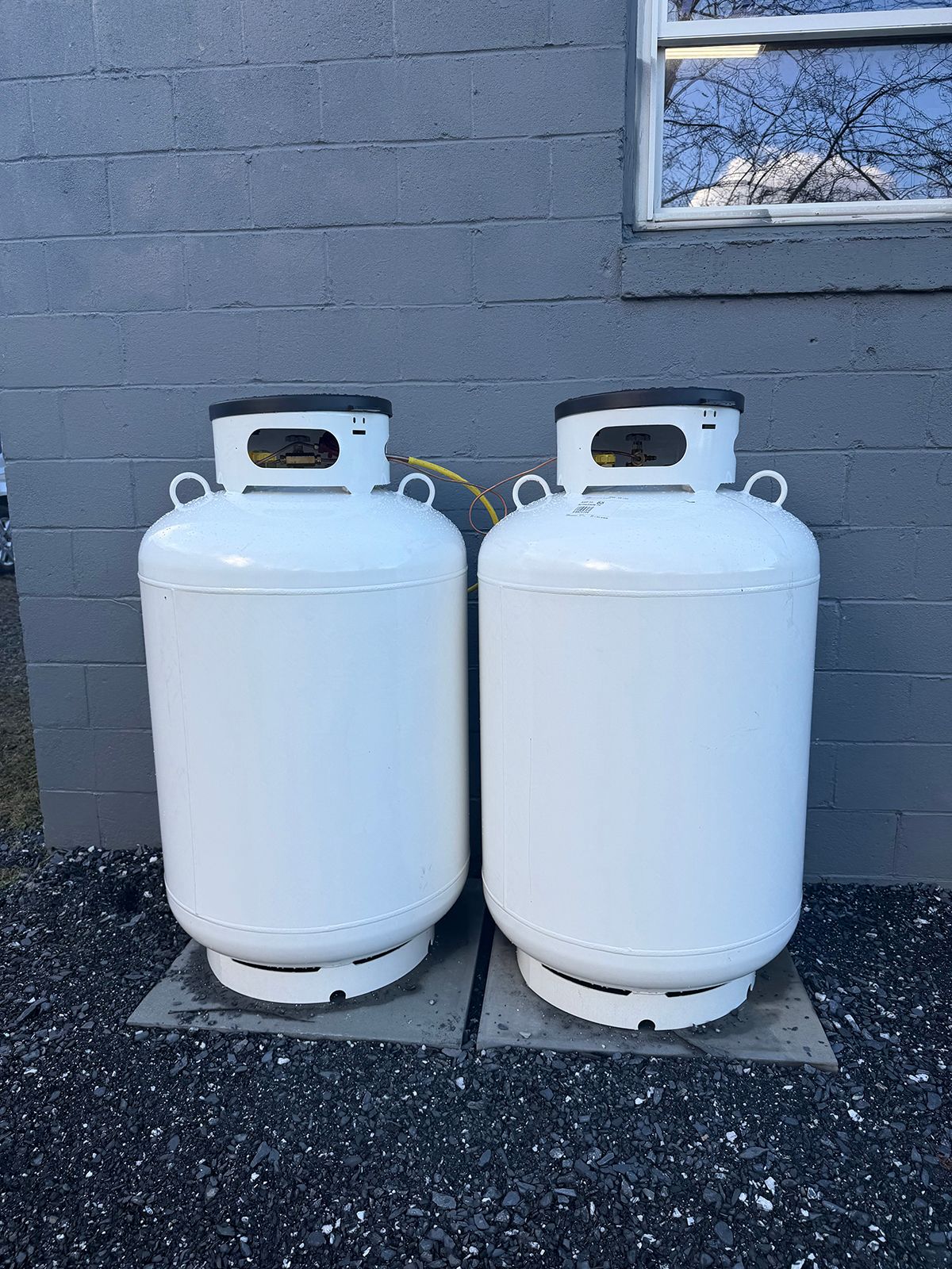 Two white propane tanks standing side-by-side on concrete pads against a gray brick wall.